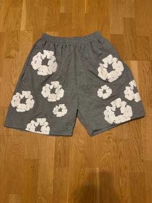 Grå denim tears shorts med vita blommor - Säljer ett par grå shorts med elastisk midja och stora vita blommönster över hela plagget. Shortsen har en avslappnad passform och är perfekta för varma dagar. Tillverkade i mjukt material som känns skönt mot huden.