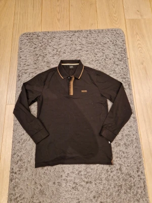 Svart långärmad skjorta från BOSS - Snygg svart långärmad skjorta från BOSS med bruna detaljer på krage och knappslå. Skjortan har klassisk krage, diskret BOSS-logga på bröstet och kontrastfärgade ränder på insidan av kragen. Perfekt för en stilren look.