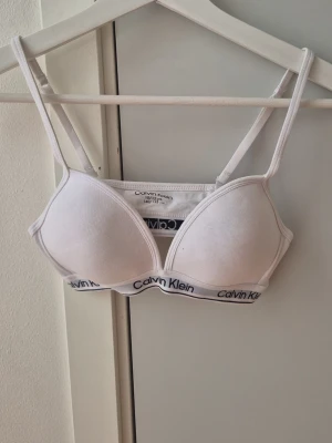 Vit Calvin Klein bh med tunna band - Snygg vit bh från Calvin Klein med tunna axelband. Elastiskt band under bysten med logotypen tryckt runtom. Klassisk och enkel design i mjukt material som ger bra stöd och komfort.
