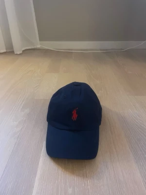Mörkblå keps från Polo Ralph Lauren - Snygg mörkblå keps från Polo Ralph Lauren med klassisk röd broderad logga framtill. Kepsen har böjd skärm och justerbar rem baktill. Tillverkad i bomull för en bekväm och stilren look.