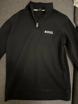 Svart halfzip tröja från BOSS - Snygg svart tröja från BOSS med halv dragkedja och hög krage. Klassisk logga på bröstet och stilren design. Perfekt till jeans eller chinos för en clean look.