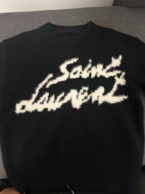 Svart stickad Saint Laurent tröja - Snygg svart stickad tröja från Saint Laurent med vit, fluffig logotyp framtill i grafisk stil. Tröjan har rund halsringning, långa ärmar och ribbade muddar vid ärmslut och nederkant. Perfekt för dig som gillar streetstyle och exklusiva märken.