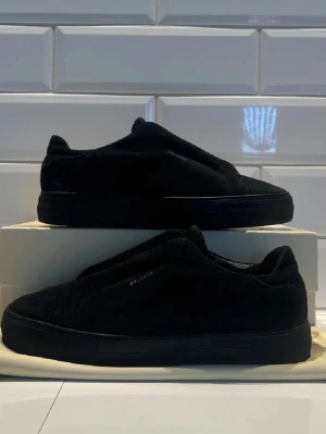 Axel arigatos - Stilrena svarta sneakers i mocka med låg profil och minimalistisk design. Skorna har en helsvart sula och ovandel, samt diskreta detaljer på sidan. Perfekta för dig som gillar en clean och modern look.