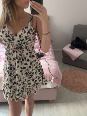 Blommig kort sommarklänning - Söt kort klänning med blommigt mönster i blått och beige på vit botten. Klänningen har v-ringning, smala axelband och volangkant nertill. Perfekt för varma dagar och har en lätt och luftig känsla.