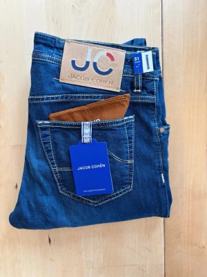 Jacob Cohën Ny - Snygga blå jeans från Jacob Cohën med klassisk femficksmodell och kontrastsömmar. Jeansen har ett stort JC-läderpatch bak och detaljer som etikett och ficknäsduk. Tillverkade i mjukt denimtyg för en bekväm passform.