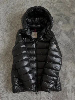 Svart dunjacka från Moncler - Snygg svart dunjacka från Moncler med glansig finish och klassisk Moncler-logga på ärmen. Jackan har huva, dragkedja framtill och quiltad design som ger en riktigt modern look. Perfekt för kalla vinterdagar och riktigt varm tack vare dunfyllningen.