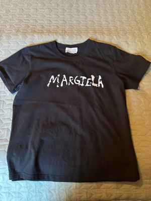 Svart Margiela t-shirt med tryck - Svart t-shirt från Maison Margiela med vitt tryck på bröstet där det står 'Margiela' i en unik, handskriven stil. Klassisk rund hals och korta ärmar. Tillverkad i mjuk bomull som känns skön mot huden. Perfekt för dig som gillar streetwear och designerplagg.