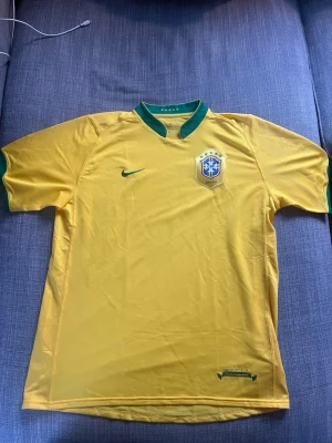 Brasilien fotbollströja Nike gul - Gul Brasilien fotbollströja från Nike med gröna detaljer vid krage och ärmslut. Tröjan har korta ärmar, broderad Nike-logga och Brasilien-emblem på bröstet. Perfekt för match eller träning. Tillverkad i lätt och ventilerande material.