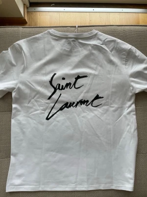 Vit Saint Laurent t-shirt med tryck - Vit t-shirt från Saint Laurent med svart logotyptryck både fram och bak. Klassisk rund hals och korta ärmar. Tillverkad i mjuk bomull för en skön och stilren look.