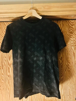 Hugo Boss T-Shirt - Säljer en svart t-shirt med ett diskret geometriskt mönster i samma ton. T-shirten har rund halsringning och korta ärmar. Materialet ser ut att vara mjukt och bekvämt, perfekt för en avslappnad stil.