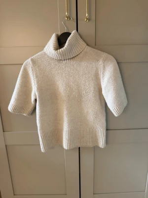 Vit stickad polotröja med kort ärm - Säljer en krämvit stickad polotröja med korta ärmar och bred ribbstickad kant vid hals, ärmslut och nederkant. Tröjan har en oversized polokrage och är gjord i ett mjukt, tjockt material som känns riktigt cozy. Perfekt för lager på lager och en clean look.