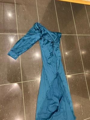 Blå långklänning med oneshoulder - Snygg blå långklänning i glansigt material med oneshoulder-design och lång ärm. Klänningen har en twistad detalj vid bysten och en slits framtill för extra stilpoäng. Perfekt för dig som vill sticka ut med en unik look.