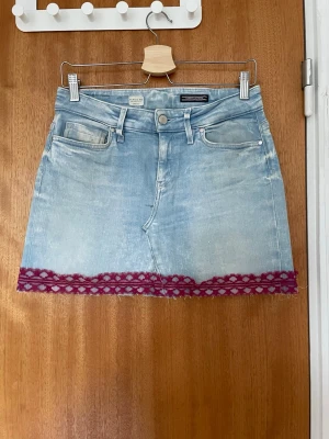 Ljusblå jeanskjol med spetsdetaljer - Kort jeanskjol i ljusblå denim med fem fickor och lila spetsdetaljer längs nederkanten och på ena bakfickan. Kjolen har klassisk knapp- och dragkedjestängning samt bälteshällor. Perfekt för en trendig och avslappnad look. Pris kan förhandlas skickar fler bilder vid förfrågan.
