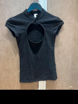 Svart topp med cut out-detalj - Snygg svart topp med korta ärmar och en stor rund cut out-detalj framtill. Toppen har en hög krage och är figurnära i modellen. Perfekt för dig som vill sticka ut med en unik och trendig look.