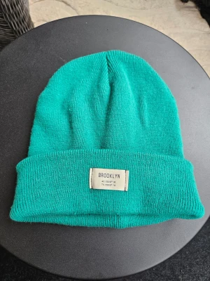 Grön mössa med Brooklyn-etikett Gina Tricot - Snygg grön stickad mössa med uppvikt kant och en vit etikett framtill där det står 'BROOKLYN' samt koordinater. Perfekt accessoar för att hålla värmen och addera lite färg till din outfit.