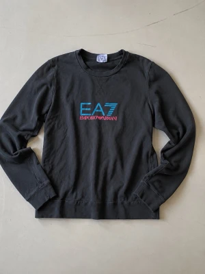 EA7 Emporio Armani Retro Logo Sweatshirt - 🖤 EA7 Emporio Armani sweatshirt med retro logotyp i blått & rosa Snyggt lätt faded svart med logo både fram och bak Mjuk och tunn sweatshirt – perfekt för en 00s/Y2K sportswear look Storleksetiketten är bortklippt, se exakta mått på bilderna Fint begagnat skick med naturliga tecken på användning Storlek: M/L, men passar som S om man vill ha lite oversize Nypris för liknande EA7-sweatshirts idag ca 1 200–1 500 kr ✨