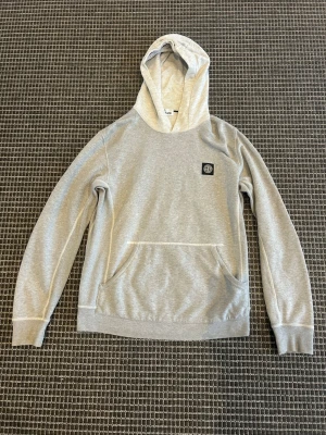 Grå hoodie från Stone Island - Snygg grå hoodie från Stone Island med klassisk logopatch på bröstet och stor känguruficka framtill. Tillverkad i mjuk bomull och har en skön huva. Perfekt för dig som gillar streetwear och vill ha en stilren look.