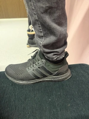 Svarta Adidas Ultraboost sneakers - Säljer ett par svarta Adidas Ultraboost sneakers med stickad ovandel och klassiska tre ränder på sidan. Skorna har en tjock, responsiv sula och snörning framtill. Perfekta för dig som gillar sportig och stilren look.