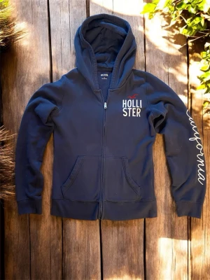 Hollister hoodie  - Mörkblå hoodie från Hollister med dragkedja framtill, broderad logga och text på bröstet samt 'California' tryckt längs ena ärmen. Utrustad med huva och kängurufickor. Perfekt för en avslappnad och cool stil.