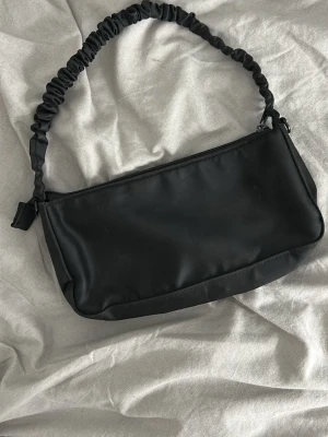 Svart axelväska med rynkat band - Stilren svart handväska i rektangulär form med ett rynkat axelband och dragkedja upptill. Väskan har en minimalistisk look och är tillverkad i ett slätt, matt material som ger en modern känsla.