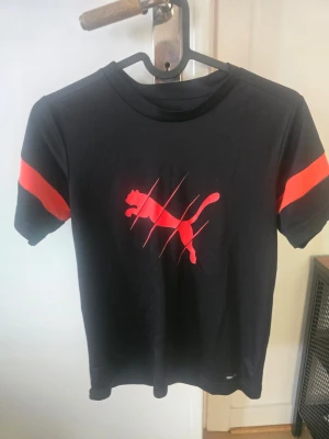 Svart Puma t-shirt med röd logga - Snygg svart t-shirt från Puma med röd logga och coolt klösmönster på bröstet.  Storlek oklart, uppskattar till 122-134.