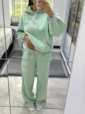 Mintgrönt loungewear-set med hoodie - Mysigt loungewear-set i mintgrönt med oversized hoodie och matchande byxor. Hoodien har magficka och broderad detalj på bröstet, byxorna har resår i midjan och samma broderi på benet. Perfekt för chill dagar hemma. Har en till två fläckar, men det går bort i tvätten 