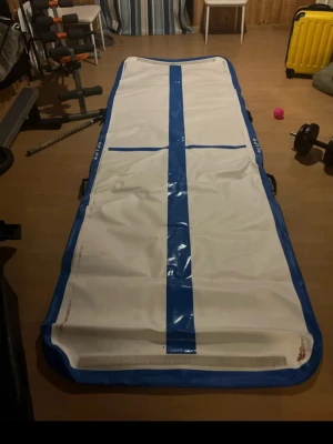 Airtrack matta blå och vit - Säljer en airtrack matta i vitt med blå kanter och detaljer. Perfekt för gymnastik, träning eller akrobatik hemma. Mattan har bärhandtag på sidorna och är tillverkad i slitstark plast. Enkel att rulla ihop och förvara.