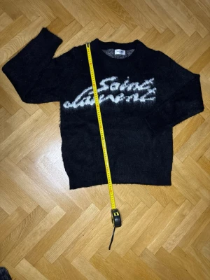 Svart Laurent stickad tröja - Säljer en svart stickad tröja från Laurent med vit, fluffig logga framtill. Tröjan har rund halsringning och lång ärm. Materialet ser ut att vara mjukt och lite luddigt, perfekt för dig som gillar en trendig och bekväm look.