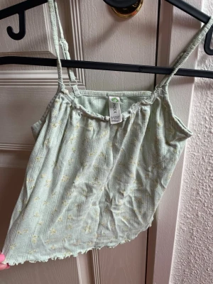 Ljusgrön blommig linnetopp, storlek  134 - Söt ljusgrön linnetopp med små blommor och tunna axelband. Toppen har en lätt ribbad struktur och vågig kant nertill. Perfekt för varma dagar och enkel att matcha med jeans eller kjol.