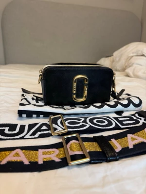 Svart Marc Jacobs Snapshot handväska - Start priset är 1500kr. Priser går att diskuteras. Säljer en svart Marc Jacobs Snapshot handväska i läder med guldiga metalldetaljer och dubbla dragkedjor. Väskan har en bred, avtagbar axelrem med text och glitterdetaljer i guld och rosa. Perfekt för dig som vill ha en trendig och kompakt väska.