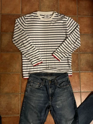 Kronstadt - Vit och svart randig långärmad topp - (Vintage) Vit långärmad tröja med svarta horisontella ränder och röda detaljer vid ärmslut och nederkant. Rund halsringning och knappdetalj på ena axeln. Perfekt för en stilren och avslappnad look.