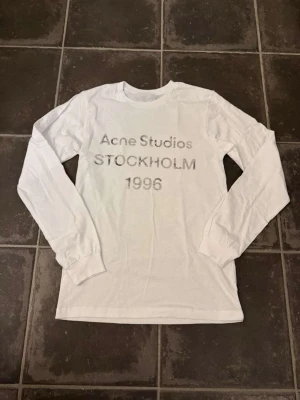 Vit långärmad tröja Acne Studios - Vit långärmad tröja från Acne Studios med tryck på bröstet där det står 'Acne Studios STOCKHOLM 1996'. Klassisk rund halsringning och rak passform. Perfekt för dig som gillar stilrena och tidlösa plagg. Acne är lite oversize men sitter som en normal M