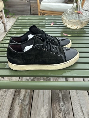 Svarta Lanvin - Snygga svarta sneakers med ovandel i mocka och tåparti i blankt lack. Skorna har svarta skosnören, metallöljetter och vit sula med retrovibbar. Perfekta för dig som gillar en stilren men ändå unik look.