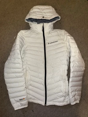 Vit pufferjacka från Peak Performance - Snygg vit pufferjacka från Peak Performance med huva och svart dragkedja. Jackan har diskreta loggor på bröstet och ärmen samt quiltad design. Perfekt för kalla vinterdagar och riktigt skön att bära tack vare det vadderade materialet.