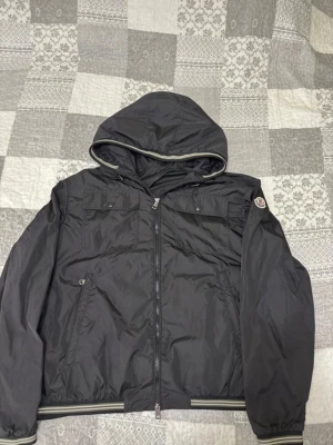 Svart vindjacka från Moncler med huva - Snygg svart vindjacka från Moncler med huva och dragkedja framtill. Jackan har två sidofickor, bröstfickor med knapp och ribbade muddar med vit rand. Moncler-logga på vänster ärm. Tillverkad i lätt och tåligt material, perfekt för blåsiga dagar.