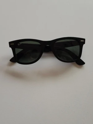 Svarta Ray-Ban Wayfarer solglasögon - Klassiska svarta Wayfarer solglasögon från Ray-Ban med bred båge och mörka glas. Snygg och tidlös design som passar till alla outfits. Ray-Ban-loggan syns tydligt på sidan och i hörnet av glaset. Perfekta för soliga dagar.