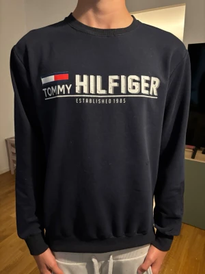 Mörkblå Tommy Hilfiger sweatshirt - Mörkblå sweatshirt från Tommy Hilfiger med stort tryck på bröstet och klassisk logga i vitt, rött och blått. Tröjan har rund halsringning, långa ärmar och ribbade muddar vid ärmslut och nederkant. Perfekt för en avslappnad och stilren look.