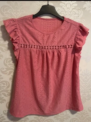 Rosa blus med volangärmar - Söt rosa blus i broderad bomull med volangdetaljer på ärmarna och rund halsringning. Blusen har ett dekorativt hålmönster och en fin spetsdetalj över bröstet. Perfekt för dig som gillar romantisk och feminin stil.