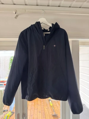 gant jacka  - Snygg svart vindjacka med dragkedja framtill och huva. Jackan har en minimalistisk design och liten logga på bröstet. Perfekt för blåsiga dagar och enkel att matcha med olika outfits. Nypris 2800kr 