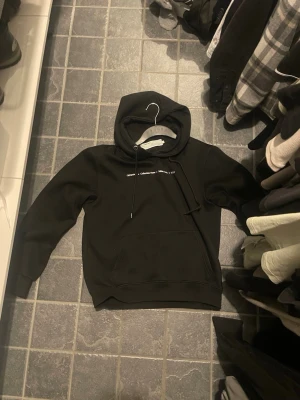 Svart hoodie  - Säljer en svart hoodie med Off-White med vit text på bröstet. Hoodien har klassisk huva med dragsko och en stor magficka framtill. Perfekt för dig som gillar streetwear och vill ha något stilrent men ändå trendigt.