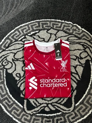 Liverpool matchtröja röd Adidas - Säljer en röd Liverpool matchtröja från Adidas med vita detaljer och klubbmärke på bröstet. Tröjan har vita ränder på axlarna, tryckt sponsorlogga och ett modernt mönster över hela tyget. Perfekt för dig som älskar fotboll och vill stötta LFC.