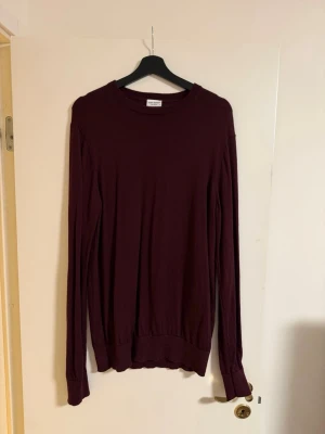 John Henric Tröja - En enkel och stilren mörkröd långärmad tröja med rund halsringning. Tröjan har ribbade muddar vid ärmslut och nederkant, och är tillverkad i ett mjukt, tunt material som känns skönt mot huden. Perfekt att matcha med jeans eller kjol.
