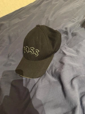 Svart keps med BOSS-logga - Snygg svart keps från BOSS med broderad vit logga framtill. Klassisk böjd skärm och justerbar passform. Tillverkad i ett strukturerat tyg som ger en sportig och stilren look.