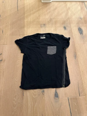 Svart t-shirt med randig ficka och dragkedja - Mörk blå t-shirt från Pelle p. T shirten är vintedge och går inte att köpa längre. Den har korta ärmar, randig bröstficka och dekorativa ränder längs sidorna. T-shirten har en avslappnad passform och är tillverkad i mjukt bomullstyg.