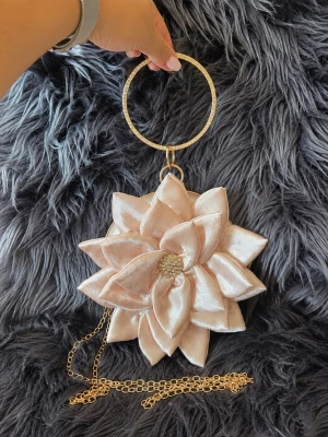 Blomformad festväska clutch puderrosa champagne strass - 🌸 Unik blomformad festväska/clutch i champagne/puderrosa satin med 3D-kronblad, guldfärgade detaljer och glittrande strass.  Avtagbar kedja medföljer så väskan kan bäras som clutch eller över axeln. Perfekt till fest, bröllop och nyår.  📏 Ca 15,5 × 15,5 × 5,5 cm Kedja ca 120 cm  🧵 Satin och metall  ✨ Endast använd några timmar vid ett tillfälle och i mycket fint skick.