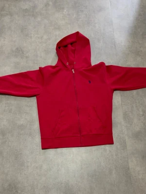 Röd hoodie med dragkedja - Snygg röd hoodie med dragkedja framtill och två fickor. Klassisk huva och ett diskret blått broderat märke på bröstet. Perfekt för dig som gillar en enkel men stilren look. Mjuk och skön bomullsmix som passar året runt. Har tvättat den för mycket därför är sista bilden så. 