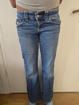 Blå bootcut jeans med låg midja - Säljer ett par blå bootcut jeans med låg midja och dubbla knappar i linningen. Jeansen har klassiska fem fickor och kontrastsömmar. Perfekta för en avslappnad och trendig look. Materialet är denim i bomull.