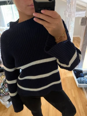 Mörkblå stickad tröja med vita ränder - Mysig mörkblå stickad tröja med breda vita ränder. Tröjan har en ribbstickad struktur, hög hals och långa ärmar med avslappnad passform. Perfekt för kyliga dagar och enkel att styla med jeans eller leggings.