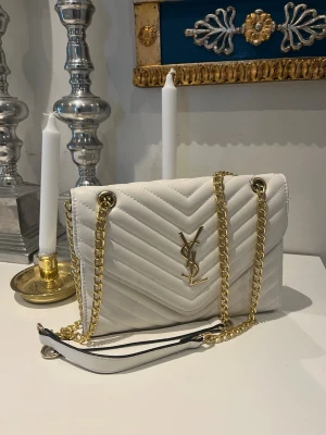 Vit kuvertväska med kedja från YSL - Snygg vit kuvertväska replika från YSL  med guldfärgade detaljer och kedjerem. Väskan är knappast använd. 25x17 cm