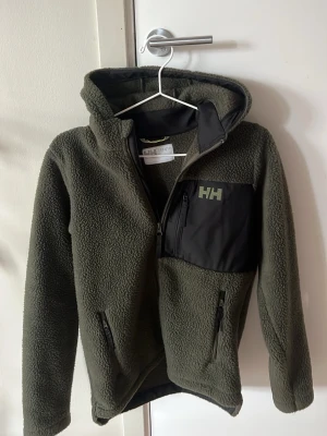 Grön fleecehoodie från Helly Hansen - Mysig grön fleecehoodie från Helly Hansen med dragkedja, huva och svart bröstficka med HH-logga. Två sidofickor med dragkedja och mjukt teddyfoder. Perfekt för kalla dagar och outdoor-äventyr. Storlek 12 år 152